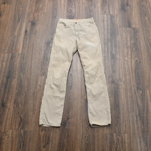 May include: Tan corduroy pants with a button and zipper closure. The pants have a classic five-pocket design and a straight leg. The corduroy fabric has a soft, textured appearance. The pants are laid flat on a wooden surface.