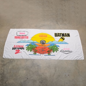 May include: White beach towel with a colorful design featuring the logos of various movies, including "Batman", "Lethal Weapon 2", "Young Einstein", and "Summer of Film Fun". The towel also features a pink Cadillac logo and a yellow sun.