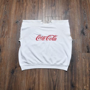 Op de afbeelding: Witte mouwloze sweatshirt met het Coca-Cola-logo in rood op de borst. Het kledingstuk heeft een ronde hals en raglanmouwen. De sweatshirt is gemaakt van een zacht materiaal, waarschijnlijk een katoenmix. De achtergrond is een houten oppervlak.