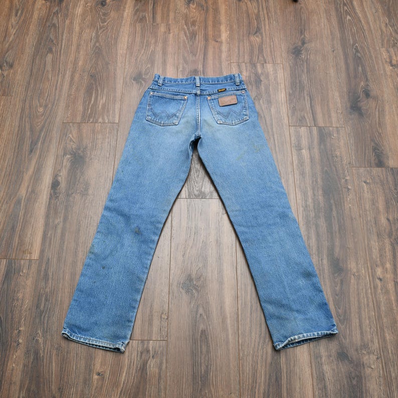 May include: Light blue denim jeans with a classic five-pocket design. The jeans feature a brown leather patch on the back and are laid out on a wooden surface. The jeans have a straight leg cut.