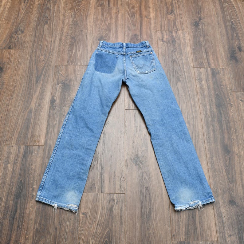 May include: Light blue denim jeans with a patch on the back pocket and distressed hems. The jeans are laid flat on a wooden surface, showcasing the back of the garment. The Wrangler logo is visible.
