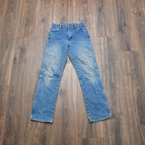 May include: Light blue denim jeans with a faded wash. The jeans have a classic five-pocket design, a button and zipper closure, and a straight leg. The jeans are laid flat on a dark wood surface.