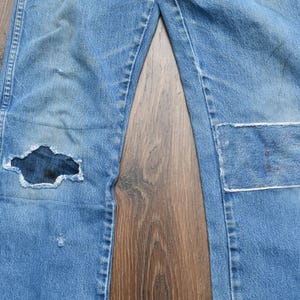May include: Light blue denim jeans with a distressed look. The jeans have a large hole in the left knee and a patch on the right knee. The jeans are laid flat on a wooden surface.
