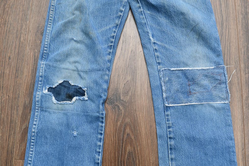 May include: Light blue denim jeans with a distressed look. The jeans have a large hole in the left knee and a patch on the right knee. The jeans are laid flat on a wooden surface.