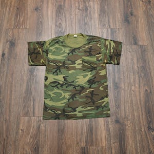 May include: A short-sleeved t-shirt featuring a classic woodland camouflage pattern in shades of olive green, brown, and black. The shirt includes a single chest pocket on the left side and a crew neckline with a solid olive green collar trim.