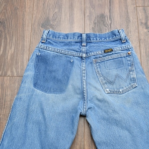 May include: Light blue denim jeans with a faded wash and a Wrangler label on the back pocket. The jeans have a classic five-pocket design and a button and zipper closure. The back pockets feature the iconic Wrangler "W" stitching.
