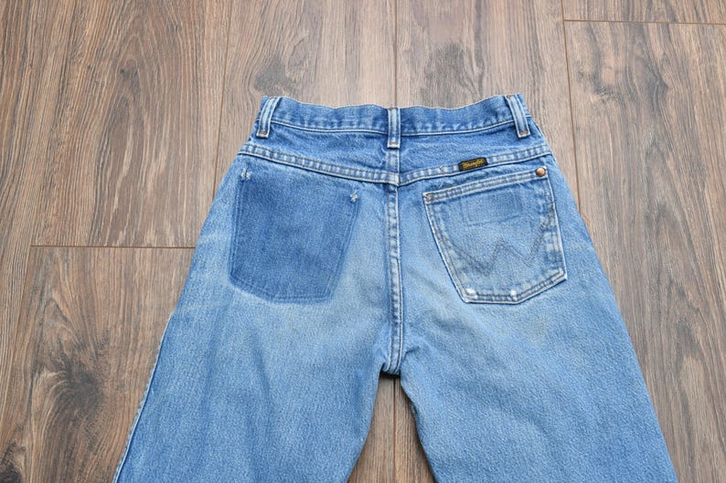 May include: Light blue denim jeans with a faded wash and a Wrangler label on the back pocket. The jeans have a classic five-pocket design and a button and zipper closure. The back pockets feature the iconic Wrangler "W" stitching.