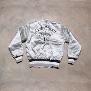以下が含まれることがあります： 黒と白のストライプの袖口とウエストバンドが付いたシルバーサテンのボンバージャケット。ジャケットの背面には、「Eddie Rabbitt Horizon」という刺繍のデザインが施されています。