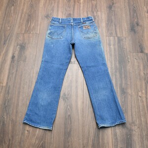 May include: Blue denim jeans with the Wrangler logo on the back pockets. The jeans have a classic five-pocket design and a straight leg fit. The denim has a faded appearance.