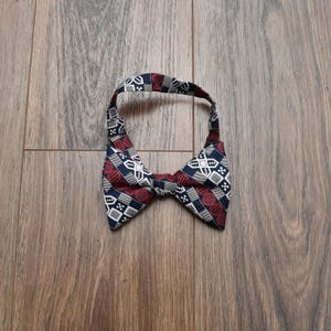 May include: A patterned bow tie with a repeating design of white, red, and navy blue shapes. The bow tie is made of fabric and is laying on a wooden surface. The bow tie is a formal accessory.