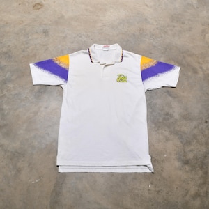 以下が含まれることがあります： 黄色と紫の斑点模様が袖に入った白い半袖ポロシャツ。シャツの前面には「Jazz」と書かれた緑と黄色のロゴがあります。