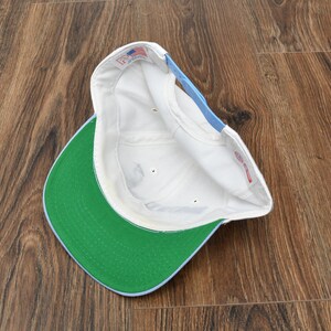 Puede incluir: Una gorra de b&eacute;isbol blanca con visera verde y un acento azul claro en la parte trasera. El interior de la gorra es blanco y la visera es de color verde brillante. La gorra est&aacute; sobre una superficie de madera.