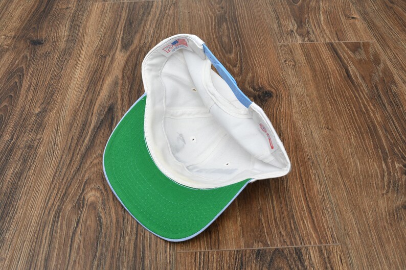 Puede incluir: Una gorra de b&eacute;isbol blanca con visera verde y un acento azul claro en la parte trasera. El interior de la gorra es blanco y la visera es de color verde brillante. La gorra est&aacute; sobre una superficie de madera.