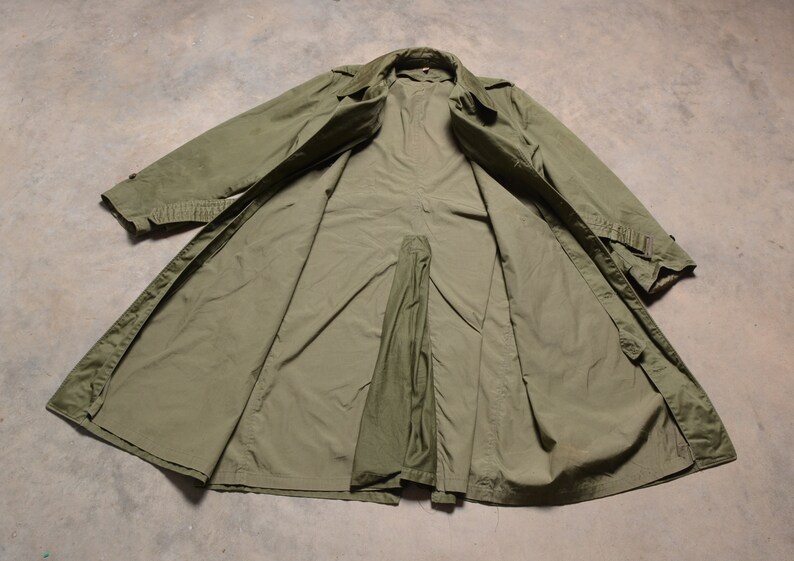 K&ouml;nnte beinhalten: Ein gr&uuml;ner Trenchcoat im Milit&auml;rstil mit Knopfleiste und G&uuml;rtel. Der Mantel liegt flach und offen und zeigt das Futter.