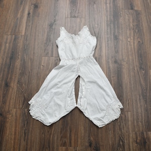 May include: White vintage jumpsuit with a sleeveless design and wide legs. The top features delicate lace detailing around the neckline and waist. The bottom of the legs have a decorative lace trim. The garment is laid flat on a wooden surface.