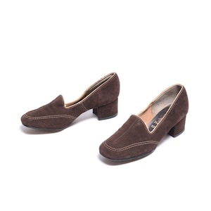 May include: A pair of brown suede heeled shoes with a low block heel. The shoes have a pointed toe and a stitched detail around the toe and heel.