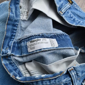 Puede incluir: Primer plano de unos vaqueros Wrangler azul claro. Los vaqueros est&aacute;n doblados, revelando el interior y una etiqueta blanca con el logotipo de Wrangler e informaci&oacute;n de tallas. La mezclilla tiene algunas zonas desgastadas.