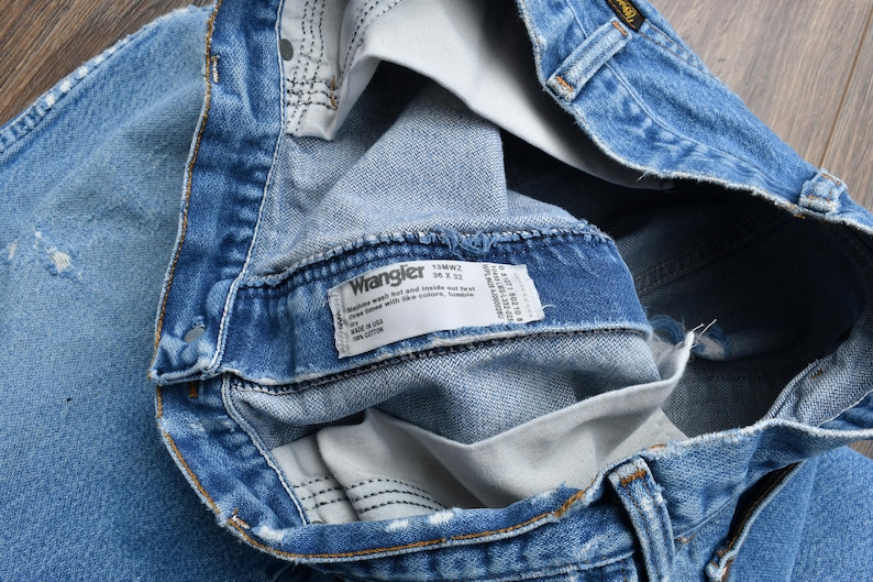 Puede incluir: Primer plano de unos vaqueros Wrangler azul claro. Los vaqueros est&aacute;n doblados, revelando el interior y una etiqueta blanca con el logotipo de Wrangler e informaci&oacute;n de tallas. La mezclilla tiene algunas zonas desgastadas.