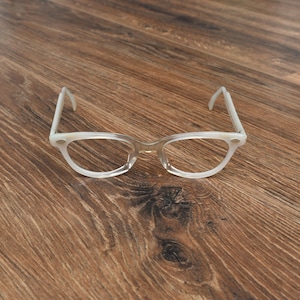 May include: A pair of vintage-style eyeglasses with a light, translucent frame. The glasses have a classic cat-eye shape with rounded lenses and white temples. The frames are set against a wooden background.