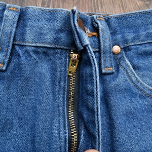 May include: Close-up of blue denim jeans with a brass zipper and copper rivets. The jeans have a classic five-pocket design with orange stitching. The fabric is a medium wash denim.