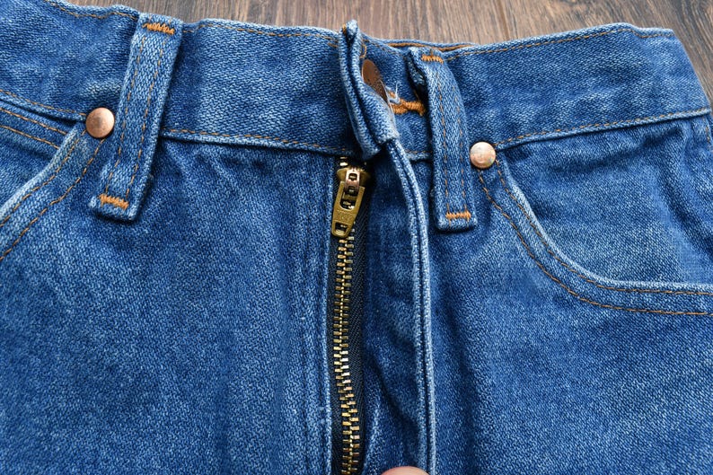 May include: Close-up of blue denim jeans with a brass zipper and copper rivets. The jeans have a classic five-pocket design with orange stitching. The fabric is a medium wash denim.