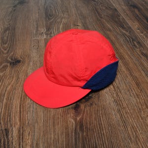 May include: A red baseball cap with a curved brim and a navy blue ear covering. The cap is made of a lightweight material and is designed to protect the ears from the cold.