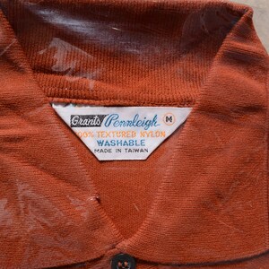 Op de afbeelding: Een bruin poloshirt met een wit label waarop staat "Grants Pennleigh 100% Textured Nylon Washable Made in Taiwan" en een maatlabel "M".