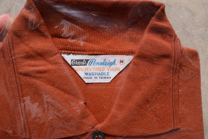 Op de afbeelding: Een bruin poloshirt met een wit label waarop staat "Grants Pennleigh 100% Textured Nylon Washable Made in Taiwan" en een maatlabel "M".