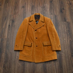May include: A mustard yellow corduroy jacket featuring a notched collar with brown trim, three front buttons, two large patch pockets with brown accents, and buttoned cuffs. The interior label displays the text "MARLBORO" in white lettering.