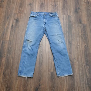Puede incluir: Vaqueros de mezclilla azul claro con un dise&ntilde;o cl&aacute;sico de cinco bolsillos. Los vaqueros muestran signos de desgaste, incluyendo &aacute;reas desgastadas y parches descoloridos. Los pantalones est&aacute;n colocados sobre una superficie de madera.