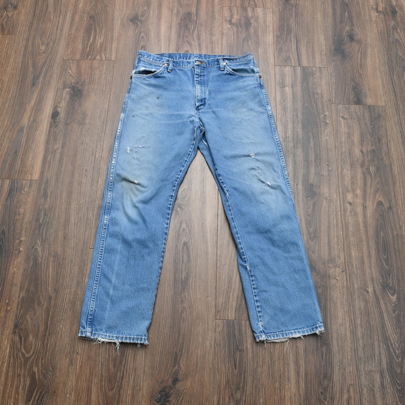 Puede incluir: Vaqueros de mezclilla azul claro con un dise&ntilde;o cl&aacute;sico de cinco bolsillos. Los vaqueros muestran signos de desgaste, incluyendo &aacute;reas desgastadas y parches descoloridos. Los pantalones est&aacute;n colocados sobre una superficie de madera.