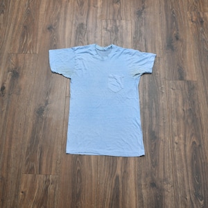 May include: A light blue short-sleeved t-shirt featuring a crew neckline and a single chest pocket on the left side. The garment displays visible discolouration under the arm areas, consistent with wear, and is constructed from a plain fabric.