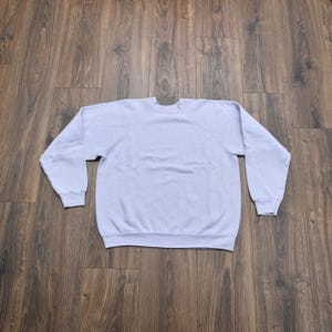 May include: A light purple sweatshirt with long sleeves. The crew neck sweatshirt is made of a soft material. The garment is laid flat on a wooden surface.