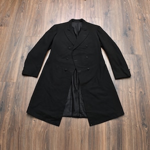 May include: A black double-breasted coat featuring a structured collar, long sleeves with buttoned cuffs, and a smooth interior lining. The garment has a series of matching black buttons down the front and a knee-length hemline with a tailored fit.