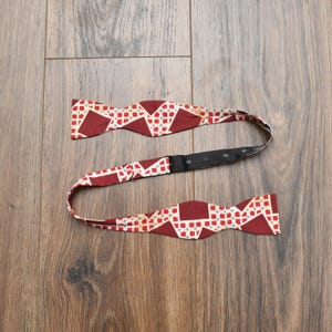 May include: A burgundy bow tie with a geometric pattern of white and gray shapes. The bow tie is made of fabric and has an adjustable black strap. The bow tie is laying on a wooden surface.