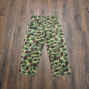 May include: Camouflage pattern pants in shades of green, brown, and tan. The pants have a relaxed fit with a button and zipper closure. The fabric appears to be a durable cotton blend. The pants are designed for outdoor activities or casual wear.