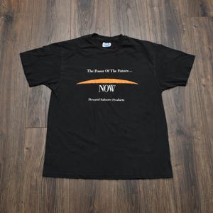 Può includere: Una t-shirt nera a maniche corte con testo bianco che recita The Power Of The Future... NOW Personal Software Products, con un elemento grafico arancione curvo posizionato tra le due righe di testo sulla parte anteriore.
