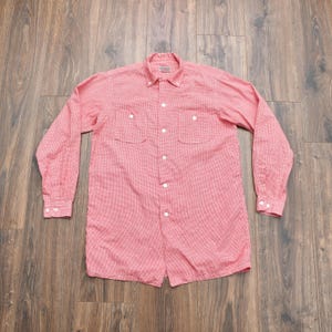 May include: A long-sleeved button-down shirt in a light red and white micro-check pattern. The shirt features a button-down collar, two chest pockets with buttoned flaps, white buttons down the front placket, and buttoned cuffs on the long sleeves.