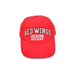 May include: A red baseball cap with the text "RED WINGS" in white and black embroidery. Below the text is "CCM HOCKEY" in white. The cap is a classic six-panel design, suitable for sports fans.