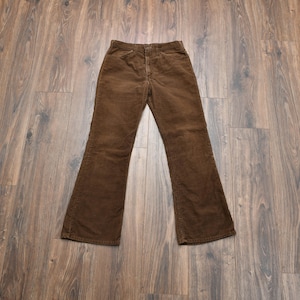 May include: A pair of dark brown corduroy pants featuring a high-waisted design, a button and zipper fly closure, two front pockets, and a slightly flared leg silhouette. The fabric has a distinct vertical ribbed texture throughout the garment.