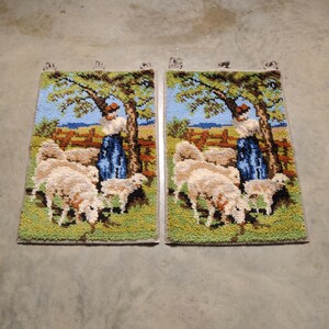 May include: Two framed needlepoint wall hangings with a pastoral scene. Each piece features a woman in a blue dress standing near a tree with sheep in a field. The background is a blue sky with white clouds.