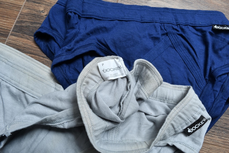 Puede incluir: Dos calzoncillos Jockey azules y grises, con el logo de Jockey visible. Los calzoncillos son de tela suave y est&aacute;n sobre una superficie de madera.