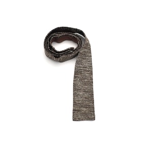 Puede incluir: Una corbata tejida marrón y beige, exhibida sobre un fondo blanco. La corbata está enrollada en la parte superior, con el extremo largo colgando. La corbata tiene una apariencia texturizada, con una mezcla de hilos marrones y beige.