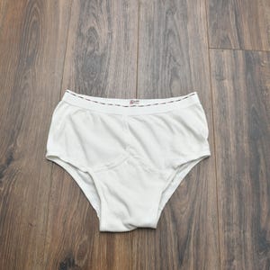 Puede incluir: Bragas blancas de dise&ntilde;o cl&aacute;sico, con un panel frontal reforzado. La cintura tiene un ribete decorativo. La ropa interior est&aacute; hecha de una tela suave y opaca. La prenda se exhibe sobre una superficie de madera.