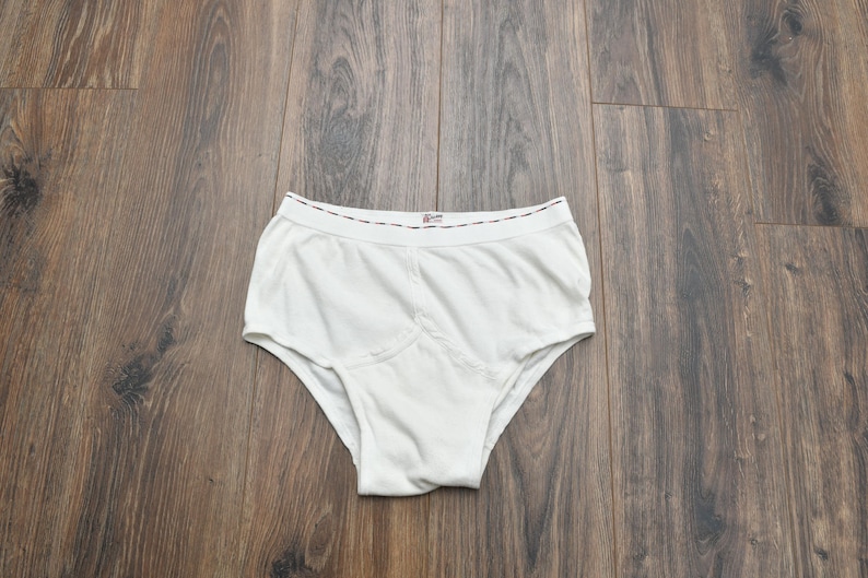 Puede incluir: Bragas blancas de dise&ntilde;o cl&aacute;sico, con un panel frontal reforzado. La cintura tiene un ribete decorativo. La ropa interior est&aacute; hecha de una tela suave y opaca. La prenda se exhibe sobre una superficie de madera.