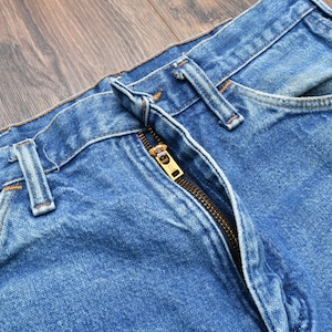 May include: Close-up of blue denim jeans with a visible zipper and belt loops. The jeans have a classic design with a faded wash, showing signs of wear. The zipper is gold-colored.