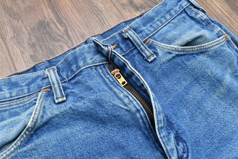 May include: Close-up of blue denim jeans with a visible zipper and belt loops. The jeans have a classic design with a faded wash, showing signs of wear. The zipper is gold-colored.
