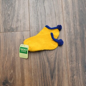 May include: A pair of bright yellow fuzzy ankle socks with a blue trim and a small blue pom-pom on each heel. A light green rectangular tag is attached to the socks, featuring the text SOCKS DESIGNED FOR STYLE & COMFORT in white and black.