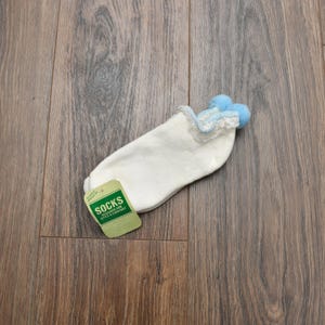 May include: White socks with a lace trim and two light blue pom-poms. The socks have a tag that reads "SOCKS DESIGNED FOR STYLE & COMFORT". The socks are on a wooden surface.