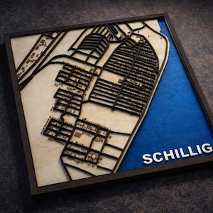 May include: A framed wooden map of Schillig, featuring a detailed laser-cut design. The map has a natural wood finish with dark brown streets and buildings, and a blue section representing the sea. The word "SCHILLIG" is displayed in white letters.
